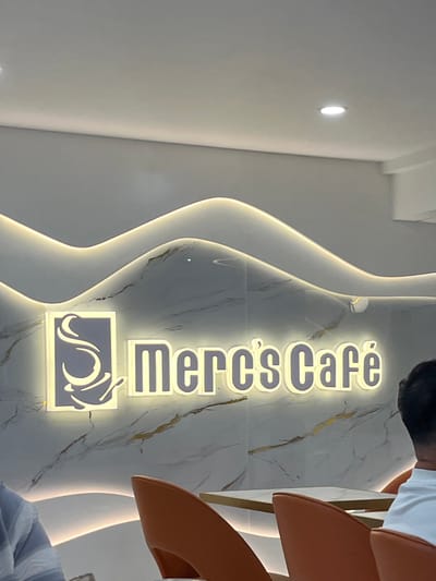 In Koronadal City, Philippines, a brightly lit sign reading "Merc's Cafe" is prominently displayed on a marble-patterned wall. The sign features a stylized logo of a coffee cup on the left, followed by the cafe's name in bold, illuminated letters. The wall behind the sign has a decorative wavy design with embedded lighting, creating an ambient glow. In the foreground, two orange chairs are visible, one partially occupied by a person seen from behind, wearing a white shirt. Another person, wearing a blue and white striped shirt, is partially visible on the left, seated and possibly looking towards the table in front of them. The overall impression is a modern and inviting cafe interior.