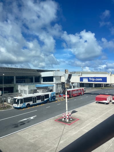 This is a daytime, outdoor, eye-level, full shot of an airport tarmac in Mai Khao, Thailand. The image is dominated by a bright blue sky with scattered white clouds. In the foreground, a concrete walkway is visible, with a diagonal shadow cast across it. The middle ground features an asphalt tarmac with white lane markings and directional arrows. Two buses are present on the tarmac: a white and blue bus on the left, and a red bus with Thai text on the right. Behind the buses, a jet bridge extends from a terminal building. A large blue banner with "Trip.com" logo is displayed on the jet bridge. Several luggage carts, one with a red cover and the number "294" visible, are parked on the right side of the tarmac. The background shows a long, low airport terminal building with dark windows. The overall scene depicts a typical airport operational area under a clear sky.
