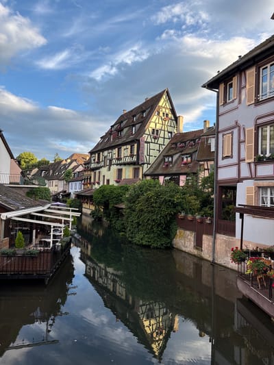 This is a photograph of a canal in Colmar, France, specifically in the area known as "La Petite Venise" (Little Venice). The scene depicts several traditional Alsatian houses lining the canal, characterized by their half-timbered facades and steeply pitched roofs. The houses are painted in various pastel colors, including yellow, pink, and light blue, with dark wooden beams.

The canal itself is calm and reflects the surrounding architecture and the cloudy blue sky. On the left side of the canal, there is a floating restaurant or cafe with tables and chairs set up outdoors. Greenery, including trees and bushes, is abundant, softening the edges of the buildings and the canal. Flower boxes adorn the windows and balconies of the houses, adding pops of red and pink to the scene.

The lighting suggests it is either early morning or late afternoon, with a mix of sunlight and cloud cover. There are no people visible in the image. The overall impression is one of a picturesque and tranquil European town.
