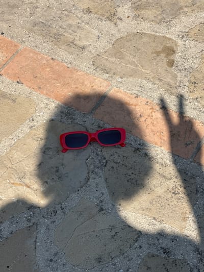 A pair of red-framed sunglasses with dark lenses lies on a stone pathway. The pathway is composed of irregular, light-colored stone slabs interspersed with reddish-brown brick sections. Shadows, cast by an unseen individual, fall across the scene. One shadow, on the right, distinctly shows a hand making a peace sign. The ground appears dry, with some minor discoloration and debris visible on the stone surfaces. The lighting suggests an outdoor setting with bright sunlight. The location is identified as Cannara, Italy.