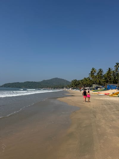 A beach landscape merges golden sand, turquoise water, and a clear blue sky. Palm trees line the edge of the beach, providing shade. Gentle waves caress the shoreline. Two individuals walk along the beach. Their reflections are seen in the wet sand. A lush, green hill rises in the background, adding to the scenic view.