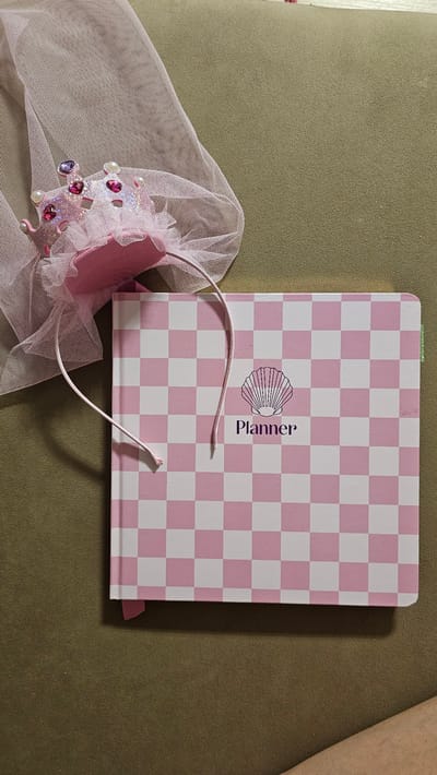 The image shows a pink and white checkered planner, lying open on a textured green surface. A delicate pink tulle veil, adorned with sparkling pink and purple jewels and pearl-like beads, is draped over the left side of the planner. The planner has a seashell graphic and the word "Planner" written vertically next to it in purple lettering. A thin, pink elastic cord is attached to the planner and loops over the veil. A small pink ribbon bookmark is tucked into the right edge of the planner. The lighting suggests it is daytime, and the setting appears to be indoors, possibly on a couch or chair. No specific location cues are visible to confirm it is in María De Huerva, Spain.