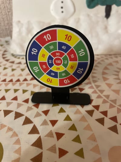 An upright, circular dartboard with a black base is centered in the frame, positioned on a white cloth surface patterned with small, randomly oriented triangles in shades of brown, pink, and green. The dartboard is divided into concentric rings and radial segments, each featuring numbers and colored sections. The outermost ring has alternating red and yellow segments, each displaying the number "10". Inside this, a blue ring shows "40" and "10". Further in, a green ring shows "30" and "10". The next ring, red, has "20" and "50". The innermost ring is red, displaying "90" and "09". At the very center is a white circle with "100". The dartboard is angled slightly towards the viewer. In the background, out of focus, is a white, fuzzy surface with a faint shadow of what appears to be a snowman and a green object resembling a Christmas tree. The lighting is ambient and appears to be from overhead. The overall scene suggests a casual indoor setting. There are no people or animals present.
