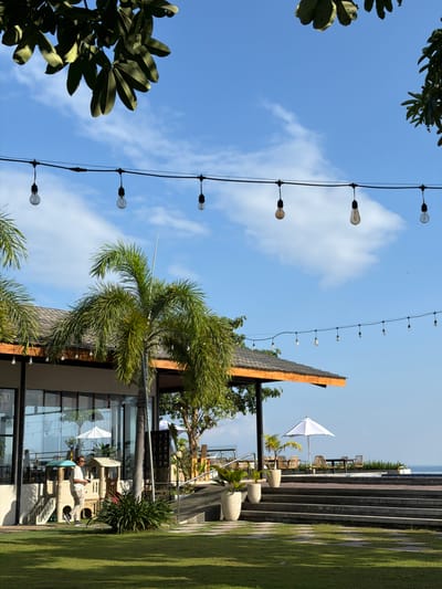 The image depicts an outdoor seating area at a resort or restaurant in Pasir Putih, Indonesia. The scene is bathed in bright sunlight under a clear blue sky with scattered white clouds. A row of string lights with exposed bulbs hangs across the upper portion of the frame, providing a festive atmosphere. In the foreground, a well-maintained green lawn leads towards a modern building with large glass windows, suggesting a dining or lounge area. Palm trees with lush green fronds are scattered throughout the scene, adding a tropical feel.
To the left, a man in a white t-shirt and light-colored pants walks across the lawn, holding a phone. Adjacent to the building, a colorful children's play structure is visible. Further back, an outdoor seating arrangement features tables and chairs under a white umbrella, set against a backdrop of the ocean in the distance. The architecture of the building incorporates dark roofing and wooden accents. The overall impression is one of a relaxed, tropical leisure destination.