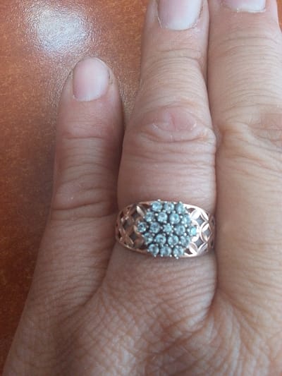 The media file shows a close-up, overhead view of a hand, specifically a ring finger adorned with a ring. The ring features a rose gold band with a patterned design on the sides. The top of the ring is encrusted with numerous small, clear stones, possibly diamonds or cubic zirconia, arranged in a cluster. The hand is resting on a textured, reddish-brown surface, which could be wood or a similar material. The lighting suggests an indoor setting, possibly with natural light as indicated by the reflections on the surface. The image focuses on the detail of the ring and the texture of the skin. There is no visible text in the image. The setting of Сарыкөл, Kazakhstan, provides a geographical context, but there are no visual cues within the image to directly confirm this location. The image does not depict any specific activity or event, other than the presentation of the ring on the finger. The mood is neutral and observational.