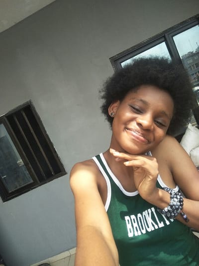 A young female subject is depicted in an outdoor selfie within an urban environment in Cotonou, Benin. She has dark, voluminous afro-textured hair and a smiling expression with her eyes partially closed. She wears a dark green tank top with white trim, bearing the word "BROOKLYN" in white letters across the chest. A small, circular, dark-colored stud earring with a lighter smiley face design is visible on her left ear (viewer's right). Her left wrist (viewer's right) is adorned with multiple black and grey beaded bracelets. Her right hand (viewer's left) is positioned with fingers gently curled, resting under her chin.

The background on the viewer's left features a plain grey concrete wall with a rectangular window, framed in dark material and fitted with vertical security bars. On the viewer's right, a larger dark-framed window or glass panel reflects city architecture, including multi-story buildings and a distant antenna or water tower, consistent with an elevated perspective within an urban area. A white object, possibly a stacked bag or cushion, is partially visible behind the subject's left shoulder. The scene is brightly illuminated by natural sunlight.