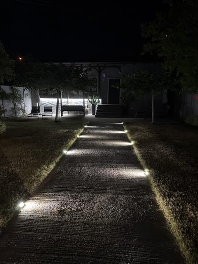 This is a nighttime, low-angle, outdoor shot of a modern residential property in Nea Moudania, Greece. The scene is illuminated by landscape lighting that casts bright pools of light on a textured, dark pathway and adjacent manicured grass. The pathway runs diagonally from the lower right to the upper left of the frame. A row of small, bright lights are embedded along the edge of the grass, lining the pathway.
To the left of the pathway and grass, there is a two-story structure with an unusual, almost abstract facade. The building appears to be made of dark material, possibly wood or a dark stone, with large windows that glow with interior light. A set of stairs leads up to a covered entrance. Green foliage, possibly trees or large bushes, frames the left side of the building and extends over parts of the structure. One large tree or bush is prominent in the upper left corner.
The overall mood is serene and modern, with the lighting creating a dramatic and inviting ambiance. The time of day is clearly night. The weather appears calm. There are no people visible in the image. The location is implied to be a private residence, likely in a coastal or garden setting, given the presence of lush greenery and the modern architectural style. No visible text is present.