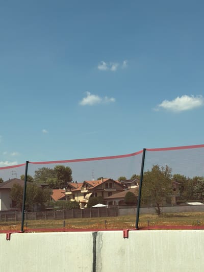 The image shows a residential area in Marnate, Italy, with several houses visible behind a construction safety net. The sky is clear blue with a few scattered white clouds. In the foreground, a concrete barrier is present, suggesting ongoing construction or road work. The safety net, which is red and black, is stretched across the scene, supported by black poles. The houses have terracotta roofs and appear to be well-maintained. Trees and greenery are also visible, providing a natural element to the urban landscape. The overall impression is one of a peaceful suburban setting undergoing development.
