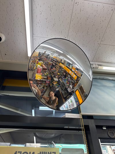 This image captures the interior of a convenience store located in Jongno-gu, South Korea. A large, circular convex security mirror is prominently positioned in the central-right, reflecting the store's environment. The ceiling, visible above and reflected within the mirror, consists of white acoustic tiles with perforations and is illuminated by long, linear fluorescent light fixtures. A white dome-shaped security camera is mounted on the ceiling in the upper-left section of the frame.
The mirror's reflection provides a wide-angle view of the store's interior, showing multiple aisles with shelves densely stocked with various packaged retail products. A refrigerated display unit is visible on the right side of the reflection, featuring a bright yellow sign with Korean text, including "고객 서비스" (Customer Service). Two individuals are clearly reflected in the mirror: a male on the lower-left, holding a smartphone and positioned as if taking the photograph, and a female in the lower-center, wearing a dark sleeveless top and looking directly towards the mirror. A golden beaded chain hangs vertically from the mirror's mounting hardware on the right. Dark structural elements and a partial, inverted yellow sign with Korean text are present along the bottom edge of the overall image.