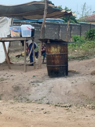 The scene depicts an outdoor, informal setting in Odo Ona, Nigeria, characterized by a dirt ground surface with sparse vegetation. In the midground, a rudimentary stall is constructed from rough wooden poles supporting a light-colored tarp roof. Beneath this roof, a wooden table holds two clear plastic buckets, one with a blue handle and the other with a red handle, containing indistinct items, along with a dark container. Behind the table, a person wearing blue trousers and a light-colored top is partially visible, standing. To their left, another individual in yellow clothing is lying on the ground.
To the right of the stall, a heavily rusted and charred metal 55-gallon drum stands on an elevated dirt mound. A metal grate rests atop the drum, indicating use for heating or cooking. Near the drum, at least one child, wearing a pink top, is partially visible. The background features a grey concrete block wall, behind which a structure with a blue roof and distant green foliage are discernible under an overcast sky. A piece of wood lies on the ground near the base of the metal drum.