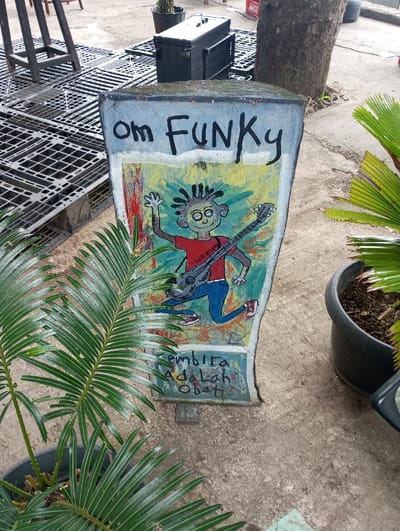 This image captures an outdoor scene in Gadog, Indonesia, focusing on a hand-painted, weathered sign standing upright on a concrete surface. The sign is roughly rectangular, wider at the top, and narrower at the base, displaying "Om FUNKy" in black letters across its upper portion. Below this text is a cartoon illustration of a grinning figure with large eyes and dreadlocks, depicted in mid-leap while playing a grey electric guitar. The figure wears a red shirt, blue pants, and red sneakers, set against a background painted with patches of yellow, green, and red. Below the illustration, the phrase "Gembira Adalah Obat!" (Joy is Medicine!) is painted in blue-green.
In the immediate foreground, to the left, is a dark potted plant with numerous long, dark green pinnate leaves, some extending across the sign. Another similar potted plant is partially visible to the right. The ground throughout the visible area is composed of uneven, light grey concrete. In the background, to the left of the sign, multiple black, grid-patterned plastic or metal pallets are arranged on the ground, with a black rectangular equipment case and a dark metal stool-like frame also present. To the right of the sign, a large tree trunk rises from the concrete. A black planter pot filled with soil is positioned near the base of this tree, and a red object, possibly a crate, is partially visible behind the tree.