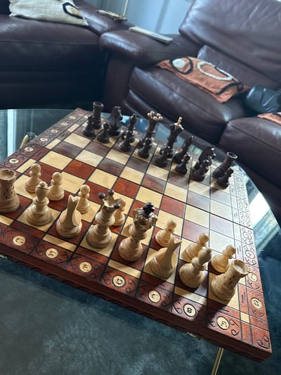 The image shows a chessboard with chess pieces set up for a game. The chessboard is made of wood, with alternating light and dark squares. The pieces are also wooden, with a classic Staunton design. The board sits on a glass table, next to a brown leather couch with decorative pillows. The surrounding environment is indistinct, not providing any forensic clues about a specific location in Palaiseau, France. There are no people visible in the image. The focus is on the chessboard and its pieces. The light is coming from the bottom left, giving the scene an average luminance.