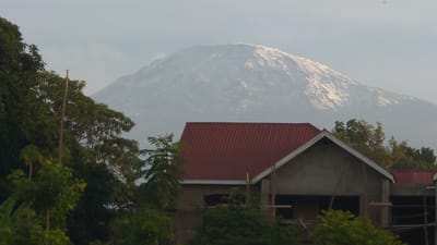 A snow-capped mountain looms in the background, partially obscured by hazy skies. In the foreground, lush greenery contrasts with a partially constructed building featuring a red roof. The building appears to be made of concrete blocks and wooden beams, hinting at an ongoing construction project. The trees in the foreground add depth to the scene.