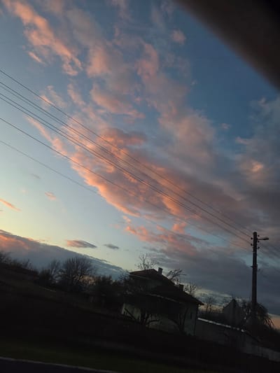 The image is a low-angle shot of a scene in Koilovtsi, Bulgaria. The focus is on a twilight sky with pink-tinged clouds. In the foreground, power lines stretch across the frame. To the right, a silhouette of a house with a chimney and bare trees is visible, along with a telephone pole. A dark, undefined shape occupies the left corner of the frame, potentially a part of a structure or object the image was taken from. The overall lighting is low, suggesting dusk or dawn.