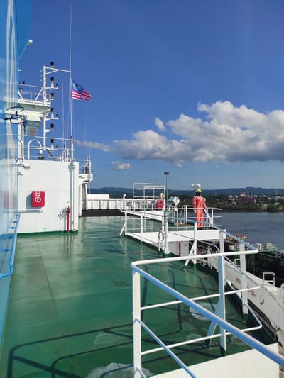 This photograph captures a broad view from the deck of a large vessel on a bright, sunny day, looking towards the starboard side and a distant coastline. The clear blue sky, adorned with fluffy white cumulus clouds, fills the upper left portion of the frame, indicating excellent weather. Below the sky, the calm, dark blue water of the sea stretches towards a green and hilly shoreline, where industrial buildings and structures are discernible along the coast.

The ship's vibrant green deck occupies the mid-ground and foreground, marked by prominent shadows of its superstructure and white railings, suggesting a strong overhead sun. Various white ship components are visible, including railings, masts, and antenna arrays. A Malaysian flag (Jalur Gemilang) proudly flutters from one of the masts, confirming the location as Kunak, Malaysia.

Further details on the ship include a red fire hose cabinet mounted on a white wall, displaying a white "F" symbol and a fire hose icon. An orange covered life raft and a red lifebuoy are also secured on the deck. Partially visible text, possibly "LIFE-SEAT" or similar, can be seen on a white panel. No people are visible in the frame. The scene conveys a sense of peaceful transit or anchoring in a coastal industrial area under clear blue skies.