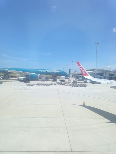 The image depicts an airport tarmac under a clear blue sky. A large, turquoise airplane with "Vietnam Airlines" written on its side is parked. Adjacent to it, a wing of another airplane, bearing the "AirAsia" logo in red and white, extends into the frame. Several service vehicles, including a fuel truck and various carts, are positioned around the Vietnam Airlines aircraft, suggesting it is undergoing servicing. In the background, an airport terminal building and a tall lighting mast are visible. The tarmac itself is paved with concrete, marked with lines, and a solitary orange traffic cone casts a long shadow. The scene is static, capturing a moment of activity at the airport. The location is identified as Bắc Cam Ranh, Vietnam.