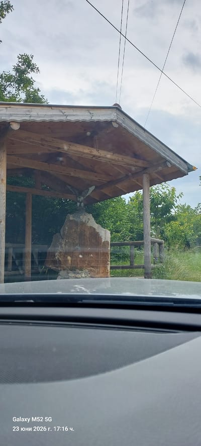This is a vertical, eye-level, slightly tilted media file showing an outdoor scene captured through a window. The primary subjects are a wooden gazebo with a gabled roof and exposed wooden beams, and a large, weathered rock formation. The gazebo appears rustic, with a decorative edge along the roofline. To the right of the gazebo, the rock stands prominently, resting on a stone base, with a wooden structure resembling a fence or railing behind it. Green foliage from trees and bushes forms the background, suggesting a natural or park-like setting.

There is no discernible activity or event taking place; it is a still outdoor scene. The lighting suggests it is daytime, likely with overcast skies given the bright, diffused light and some visible clouds. The presence of a modern vehicle window framing the scene, with a reflective surface on the right, indicates the photo was taken from inside a car.

Notable details include the texture of the rock and wood, the details of the gazebo's construction, and the lush greenery. Visible text at the bottom right corner of the image reads: "Galaxy M52 5G 23 юни 2026 г. 17:16 ч.". This text provides information about the device used to capture the image, the date (June 23, 2026), and the time (17:16, or 5:16 PM). The location context provided is Ogradna, Bulgaria. The overall mood is peaceful and observational.