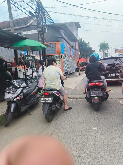 The scene takes place in the street in Kecamatan Dayeuhkolot, Indonesia. There are three motorbikes parked on the street, a black car and a white car are parked on the opposite side of the road. A man in a yellow floral shirt is sitting on a black motorbike, while a man in a black jacket is sitting on a blue motorbike behind a woman in a black dress. A woman is standing in front of the motorbike with the yellow shirt man. There is a green umbrella-covered stand on the left-hand side of the photo. The road surface appears to be asphalt. The sky is overcast with a hazy sky.