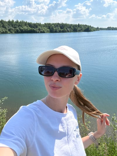 A woman stands near a body of water, taking a selfie. She is wearing a white t-shirt, a beige baseball cap, and black sunglasses. Her hair is tied back in a ponytail. The water is calm and reflects the blue sky with scattered clouds. A dense line of trees forms the backdrop on the opposite shore. The foreground shows some green vegetation and the ground appears to be sandy or earthy. The overall setting suggests an outdoor recreational area, possibly a lake or reservoir within the city of Zaosinovo, Russia.