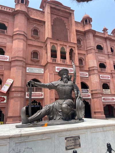 The image shows a statue of Baba Banda Singh Bahadur in front of a red sandstone building. The statue depicts him in armor, seated with a sword in his left hand and a bow and arrow in his right. He is wearing a turban and a beard. The building behind him appears to be a historical site, possibly a fort or a religious complex, with arched windows and ornate carvings. There are signs in Punjabi and English on the building, indicating it is a public space with commercial establishments. The ground in front of the statue is paved with marble. An orange flower lies on the marble base of the statue. The sky is clear and bright, suggesting it is daytime. The statue is located at the entrance of the Akal Takht in Amritsar, India.