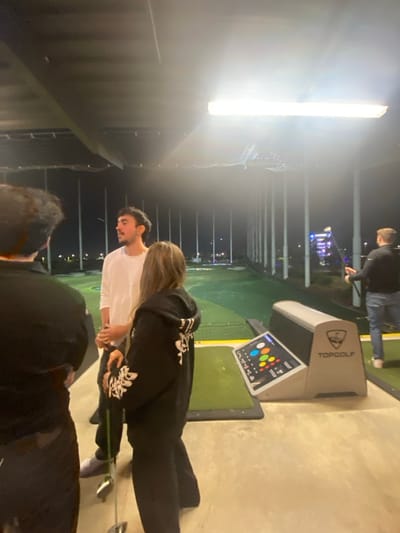 A group of people are at a Topgolf venue, likely at night. A man in a white shirt and a woman with blonde hair wearing a black hoodie stand together, possibly golfing. Another person is near the camera, partially obscured. A Topgolf monitor displaying colored circles is visible. In the background, other people golf, and the venue's infrastructure is visible with lights and barriers.