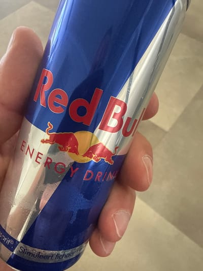 The image displays a close-up of a person's hand holding a chilled can of Red Bull energy drink. The can features its iconic design with a prominent blue section and a diagonal silver stripe. Significant condensation droplets are visible on the can's surface, indicating it is cold.
The Red Bull logo, consisting of two red bulls charging before a yellow sun disc, is clearly visible. Above this, the brand name "Red Bull" is boldly printed in red capital letters, with "ENERGY DRINK" below the logo. On the silver band at the bottom right of the can, partial text is discernible, including "Stimuleert lichaam" and "sprit®", suggesting a Dutch or Flemish language variant, likely from the phrase "Stimuleert lichaam en geest" (Stimulates body and mind).
The hand gripping the can shows visible knuckles and fingernails. The background is softly blurred and appears to be a light-colored, possibly tiled or plain wall surface, indicating an indoor setting. The activity depicted is simply holding the beverage. This scene takes place in Diekirch, Luxembourg.