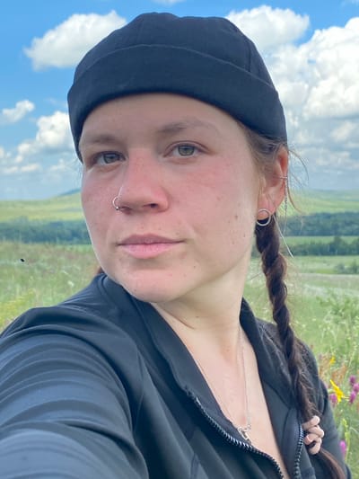 The image shows a close-up, selfie-style shot of a woman with light skin, green eyes, and light brown hair styled in a braid and worn under a black beanie. She has a nose ring and earrings. She is wearing a black zip-up jacket. The background is a landscape with green hills, scattered trees, and a blue sky with white clouds, suggesting an outdoor setting, possibly in or near the village of Ziyanchurino, Russia, given the open, rolling terrain. The focus is on the woman's face and upper body, with the landscape softly blurred.
