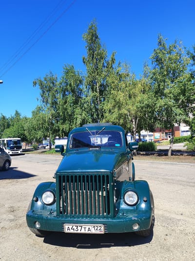 The image displays a dark green vintage truck parked on a gravel lot in Slavgorod, Russia. The truck is the primary subject, positioned centrally and viewed from the front. It features a prominent vertical grille, round headlights, and a visible license plate reading "A437TA 122 RUS". Behind the truck, a row of green deciduous trees lines a paved area where other vehicles are present, including a white bus to the left and a small car to the far left. In the background, buildings with windows and signs are visible, consistent with an urban or semi-urban setting. The sky is clear and blue, indicating fair weather. The overall scene depicts a sunny day in a Russian town.