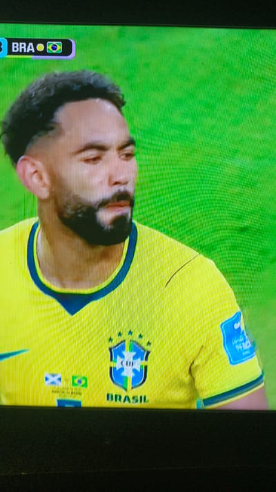 A man wearing a yellow soccer jersey with a blue collar is shown. The jersey has a crest with stars and the word "BRASIL" below it. A logo with the letters "FIFA" is visible on his right sleeve. The background is a solid green, likely a soccer field. A scoreboard graphic in the upper left corner indicates "BRA" and the Brazilian flag. This image is not from Пистамазор, Tajikistan.