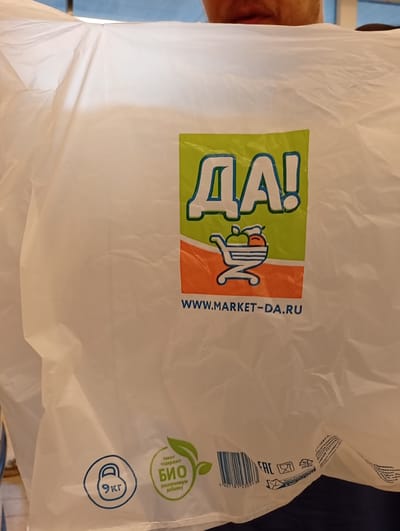 This is a close-up, slightly blurred image of a person holding a white plastic shopping bag. The bag has a large, colorful logo that reads "ДА!" in green and blue, with a stylized shopping cart graphic containing fruit. Below the logo, the website "WWW.MARKET-DA.RU" is printed.

On the lower left of the bag, there is a blue circle with a weight symbol and the text "9 кг" (9 kg). To the right of that, a green leaf emblem signifies "БИО" (BIO) and states it contains a "разлагаемую добавку" (biodegradable additive). A barcode is also visible on the bag, along with recycling symbols including "02 HDPE".

The background is out of focus but appears to be indoors, with a glimpse of a person's mouth and chin at the top of the image. The lighting suggests an indoor setting, possibly a store or a home. There are no strong cues for time of day or weather. The overall impression is of a product being displayed or used.