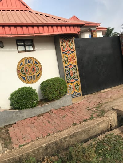 The image shows a section of the Obafemi Awolowo University Teaching Hospital complex in Oko Erin, Nigeria. A multi-story building with a coral-colored corrugated metal roof and white walls is visible. The building's design incorporates decorative elements, including a circular mosaic or relief on the wall between a window and two round bushes. A patterned, colorful frieze runs along the top of a retaining wall. A black loading platform is visible above the frieze, and the ground area includes red brick paving bordered by concrete. Plant life and vegetation growth are apparent alongside the paving. The sky is visible in the background. There are no people visible in the image.