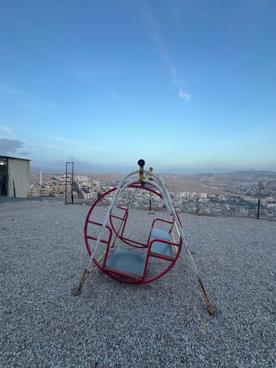 The image depicts an elevated, unpaved plot of land overlooking the city of Qudsaiya, Syria. In the foreground, a worn, red and white metal merry-go-round swing set with two light-grey seats stands on a surface covered in light-colored gravel. Rust is visible on the white painted support legs of the swing set. To the left, a small, light-colored rectangular building with a dark, open entrance is visible, adjacent to a vertical dark metal grid structure. Beyond this, a sprawling urban landscape of densely packed light-colored buildings extends across undulating terrain. A tall, slender minaret-like structure is discernible on the left side of the cityscape. The background consists of hazy, undulating hills under a clear blue sky with sparse, wispy clouds. No individuals are present in the scene. The lighting suggests either early morning or late afternoon conditions.