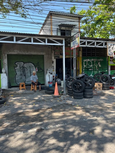 The image shows a street scene in Ngampelsari, Indonesia. A man wearing a hat and a t-shirt sits on a plastic stool outside a shop. The shop appears to be a tire repair or sales business, with stacks of tires visible in front of it. The shop has a green metal roll-up door with graffiti on it. A few other people are partially visible inside the shop. The street is paved, and there are some fallen leaves on the ground. The overall impression is that of a busy, working-class commercial area on a sunny day.