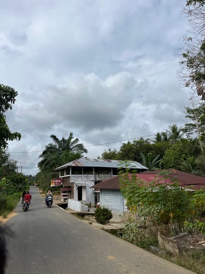 The image depicts a street scene in the city of Sukamulia, Indonesia, under a cloudy sky. A paved road runs through the center of the frame, with lush green vegetation and trees on both sides. Several houses are visible along the road. One house, in the mid-ground, features a white exterior with a balcony and a corrugated metal roof. Adjacent to it is a smaller structure with a red metal roof and light blue wooden siding. In the foreground, two motorcycles are traveling on the road. The first motorcycle, closer to the viewer, is ridden by a person wearing a red shirt. The second motorcycle, further ahead, is ridden by a person wearing a hijab and blue clothing. A few scattered pieces of debris are visible on the side of the road. The overall atmosphere is calm and rural.