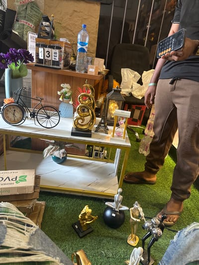 The scene depicts an interior space, likely a showroom or retail area, in Naze, Nigeria, characterized by artificial green turf flooring. A two-tiered white marble-patterned coffee table with a gold-colored metal frame occupies the central foreground.
On the top tier of the coffee table, from left to right, are: a light blue vase holding purple artificial flowers, a black metal decorative bicycle with a clock integrated into its front wheel, a light blue textured vase containing white artificial flowers, a large golden abstract spiral sculpture, a miniature black Eiffel Tower replica, a black metal lantern with an illuminated interior, and a white-framed hourglass filled with pink sand.
Behind the coffee table, a wooden cabinet or shelf unit displays various items: a perpetual calendar showing "13 FEB," a clear plastic bottle of Evian water, and a digital clock displaying "12:03." Above the cabinet, several burlap sacks labeled "FRAGILE" are stacked.
On the bottom tier of the coffee table, a potted plant with reddish-green foliage sits in a teal pot.
On the artificial turf floor in the foreground, several decorative figurines are arranged: a golden abstract figure, a silver wire figure seated on a black sphere, a silver figure playing a violin, and a black abstract figure with silver spherical elements. To the far left, stacked cardboard boxes are visible, one labeled "PVC."
Two individuals are partially visible. On the right, a person stands, wearing a dark short-sleeved shirt, brown sweatpants, and brown flip-flops. This individual holds a smartphone horizontally in their left hand, displaying the numerical value "354,000." A light green bracelet is present on their left wrist. In the bottom left, a portion of another person's leg is visible, clad in ripped blue jeans, suggesting they are seated or kneeling on the floor.
The background includes a light-colored wall and a dark office chair with white plastic bags draped over it.