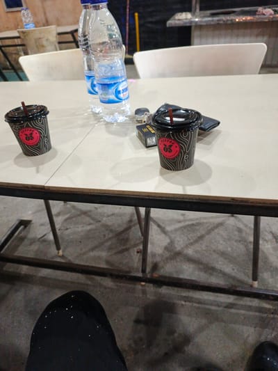 The image depicts a table at an outdoor eatery or cafe. Two disposable coffee cups with brown lids and straws are placed on the table. One cup is in the foreground on the left, and the other is to the right, slightly behind and to the right of the first. Next to the right cup, a black wallet or cardholder and a pack of cigarettes are visible. In the background, two plastic bottles of water are standing upright on the table. The table appears to be made of light-colored material, possibly tile or laminate, and is supported by a metal frame. The ground beneath the table is concrete and appears to be outdoors. The lighting suggests it might be daytime or under artificial lighting. The context of Tambonbaroh, Indonesia is inferred from the user's prompt, but there are no explicit visual cues within the image to confirm this specific location.