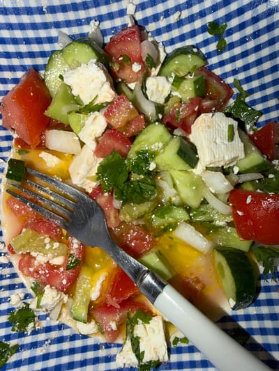 A close-up, overhead shot shows a Greek salad with tomatoes, cucumbers, feta cheese, and chopped parsley, dressed in a vinaigrette. A fork rests on the salad, its handle extending out of the frame. The salad is served on a blue and white checkered plate. The image is likely taken in Dromolaxia - Meneou Municipality, Cyprus.