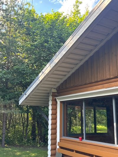 This is a close-up, low-angle shot of the exterior of a wooden house, likely in Saue, Estonia. The scene is dominated by the house's wooden siding and a large window. The siding is a warm brown color, with some areas showing a lighter, natural wood tone. The window is dark-tinted and divided into multiple panes, reflecting the surrounding greenery of trees and a blue sky with some clouds. A bright red cup is visible attached to the window frame, a small detail against the natural tones.
The architecture suggests a rustic or traditional style. The eaves of the house are visible, with decorative white scalloped trim along the lower edge. The roofline slopes upwards, with wooden planks forming the underside of the eave. The overall impression is of a sunny day, given the bright light and clear sky. The abundance of trees and the presence of what appears to be a lawn suggest a semi-rural or suburban setting. No people are visible in the image, and the focus is on the architectural details and the natural surroundings. The time of day appears to be daytime, with the sun casting light and shadows.