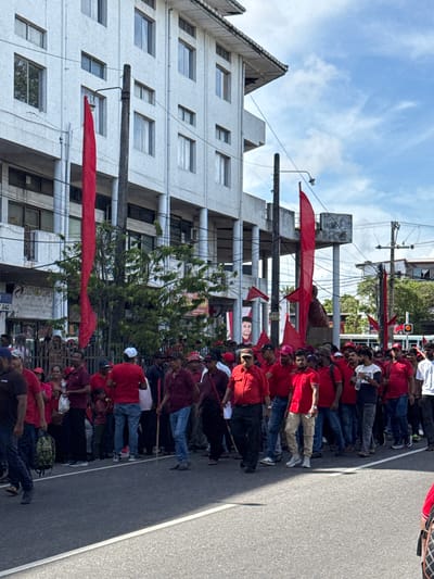 A procession of people, predominantly wearing red shirts and some with red flags, are marching down a street in Delkanda, Sri Lanka. The march is taking place on a paved road next to a multistory building. Overhead power lines are visible, as are trees lining the road. Some individuals are carrying sticks or poles. The building is white with several windows. A billboard is seen above the crowd. A large portrait is visible in the background.