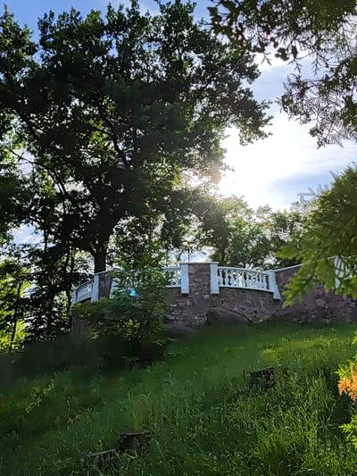 A sun-drenched outdoor scene features a stone bridge with a white balustrade, surrounded by lush green grass and dense foliage. The bridge is adorned with decorative white posts and railings, suggesting a park or garden setting. The sun's rays pierce through the leaves of a large tree, casting a dappled light across the landscape. The background reveals a bright sky with a few clouds. The image is captured in the city of Korosten, Ukraine.