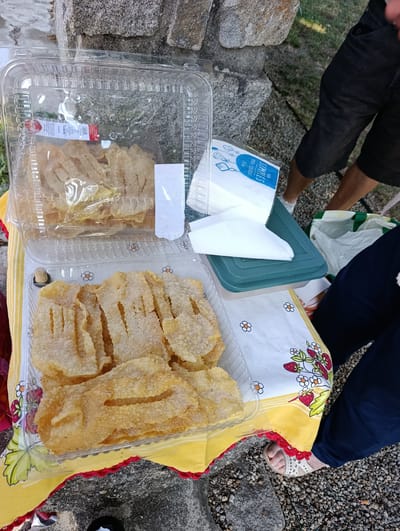 In Trofa, Portugal, a picnic setting features two transparent plastic containers of fried pastries, likely "coscorões" or a similar confection, dusted with sugar. The containers are placed on a yellow and white checkered cloth adorned with a strawberry and flower pattern. To the right of the pastries, a stack of white napkins with blue lettering, including the word "SIMPLES," is visible on a green plastic container. In the background, the lower legs and feet of at least two individuals are present. One person wears dark shorts and white shoes, while another wears dark pants and white sandals. The ground is covered in gravel and some natural debris. A large, rough stone structure is situated behind the picnic setup. The overall scene suggests an outdoor gathering where food is being shared.
