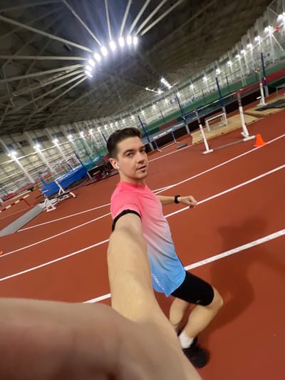 A man in athletic wear takes a selfie on an indoor track. He wears a pink and blue shirt, black shorts, and black shoes. He also has a wrist watch and a heart rate monitor on his arm. He’s standing on a red track with white lines, facing the camera. The track is in a large indoor arena with a high ceiling and bright lights. Hurdles are visible to the left of the frame.