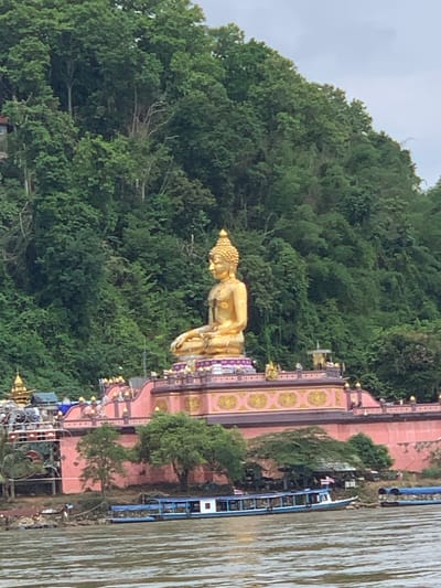 A golden Buddha statue sits prominently on a pink tiered platform against a lush green hillside in the Golden Triangle Special Economic Zone, Laos. The statue is in a meditative pose, with its hands resting on its lap. The platform is adorned with intricate gold detailing and topped with smaller decorative structures. Below the platform, a pink wall with architectural embellishments runs along the waterfront. Several trees are scattered around the base of the structure. In the foreground, a wide, muddy river flows, with a blue and white boat docked near the shore. Another smaller blue boat is visible further down the river. The sky is overcast.