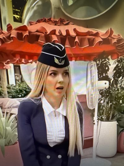 The image shows a close-up of a woman wearing a dark blue uniform with a white shirt and a dark hat with a logo. She has blonde hair and her mouth is open, suggesting she is speaking or reacting to something. The background is blurred but appears to be an indoor setting with some greenery and decorative items, including a large, ruffled, coral-colored object. The lighting is bright, and there are some rainbow-like reflections visible, possibly from the camera lens or a light source. No visible text is present in the image.