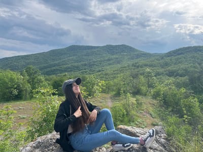 The image shows a young woman sitting on a rock, likely at a high vantage point. She is wearing a grey cap, a black jacket, a light-colored shirt, blue jeans, and sneakers. Her long, dark hair hangs down her back, and she appears to be smiling and looking upwards. The background features a landscape with lush green trees and a large hill or mountain in the distance. The sky is cloudy. This is most likely the mountainous terrain of Kamchia, Bulgaria.