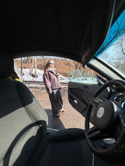 The scene is captured from within the interior of a dark-colored vehicle, specifically a Ford Mustang indicated by the emblem on the steering wheel, looking out through the open driver's side door. A female individual stands on a wet asphalt surface next to the open door. She has long dark hair and is wearing black rectangular sunglasses, a light grey hooded sweatshirt, a textured dark red/purple leather jacket, dark grey wide-leg pants with cuffed hems, and brown shoes. She is positioned facing slightly left, away from the car, with her head turned towards the vehicle's interior. In the background, beyond a metal guardrail, there is an embankment partially covered in dirty snow and slush. Bare deciduous trees are visible on the embankment under a bright blue sky. The environment suggests a cold, sunny day, likely late winter or early spring. This roadside setting is located in the city of Izmailovo, Russia.