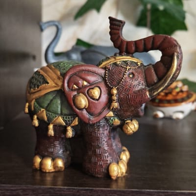 A close-up photograph shows a decorative, ceramic elephant figurine on a dark surface. The elephant has a maroon-colored body with detailed textures that mimic woven material. Gold accents highlight the feet, ear decorations, and tassels hanging from the body covering. The body covering itself is primarily green with gold edging. The elephant's trunk is raised. Another blurred, gray elephant figurine is visible in the background, along with a green plant. A small decorative container sits to the right of the elephant figurine. The environment suggests an indoor setting, possibly a shelf or display area. The image location is Novo-Nikol'skoe, Russia, but there's no indication or verification in the image itself.