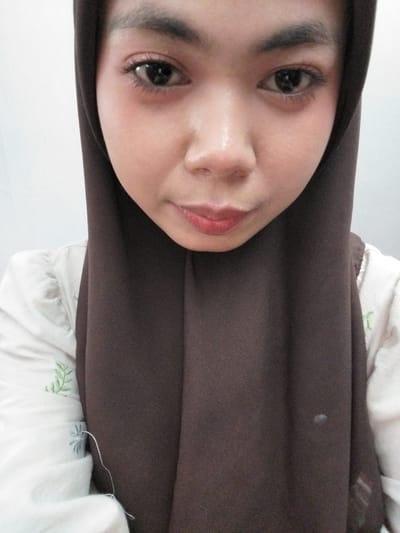 The image is a close-up, vertically oriented selfie of a young woman wearing a dark brown hijab and a white garment with a subtle green floral pattern. The woman's face is turned towards the camera, with her eyes open and looking slightly to the left of the frame. She has on dark eyeliner and mascara, and her lips are painted a reddish-pink shade. Her skin appears smooth and fair, with a subtle blush on her cheeks. The lighting suggests an indoor setting, possibly during the day, with a soft, diffused light source. A small, light-colored smudge is visible on the hijab to the right of the woman's face. The background is out of focus, but appears to be a plain white surface. The overall impression is that of a casual, personal photograph taken in a private space. The image was geotagged in Tibojong, Indonesia.
