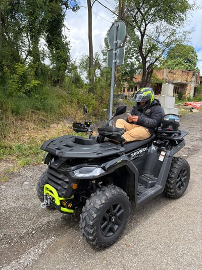 A person wearing a black jacket, tan pants, and a yellow and black helmet is seated on a black Segway ATV. Another helmet rests on the ATV's seat. The ATV is parked on a gravel road next to a patch of dry grass and a road sign. In the background, there are trees and a building that appears to be under construction or partially damaged. The location is in the city of Брусовeте, Bulgaria.
