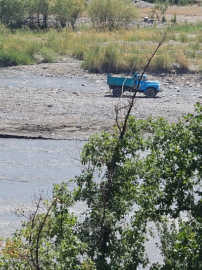 A bright blue dump truck sits parked on a rocky riverbed in Шералы, Kyrgyzstan. The truck's bed is empty, and its tires are on the gravelly ground. The river itself is a muted blue, reflecting the sky. In the foreground, the branches of a tree with green leaves obscure the view of the river. Behind the truck, tall, dry grass grows on a slight incline, leading up to a dense collection of trees in the background. The overall scene suggests a dry, arid environment.